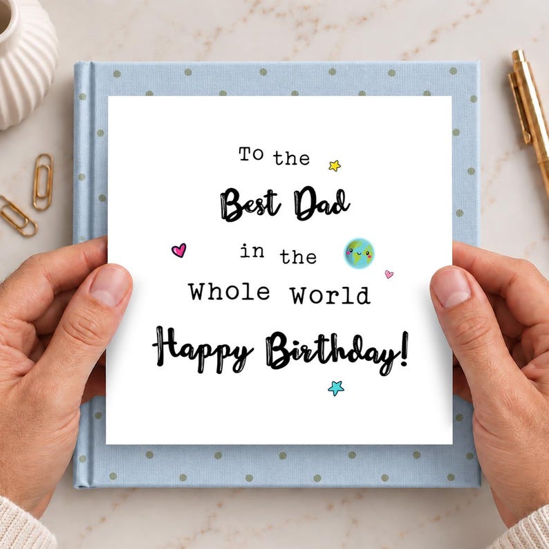 May include: A square white greeting card with black text that reads To the Best Dad in the Whole World Happy Birthday. The card features small colorful illustrations of a smiling planet Earth, two pink hearts, a yellow star, and a blue star.