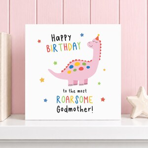 Op de afbeelding: Een vierkante witte verjaardagskaart met een roze dinosaurus die een feesthoedje draagt, omringd door kleurrijke sterren. De tekst luidt Happy Birthday to the most ROARSOME Godmother! in zwarte, rode, gele, groene en blauwe letters.