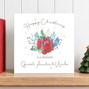 Op de afbeelding: Een vierkante wenskaart met een witte achtergrond en een aquarelillustratie van een rood cadeau, groene dennentakken en hulstbladeren. De tekst luidt Happy Christmas to a fantastic Great Auntie & Uncle.