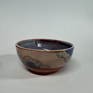 May include: A ceramic bowl with a speckled beige exterior featuring blue cloud-shaped motifs with small teal dots. The interior of the bowl has a smooth, glossy finish in a muted purple hue, and the base is a contrasting reddish-brown clay.