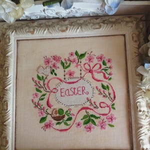 K&ouml;nnte beinhalten: Eine wei&szlig;e gerahmte Kreuzstich-Stickerei mit rosa Bl&uuml;ten, gr&uuml;nen Bl&auml;ttern und einem Band mit der Aufschrift "Easter".