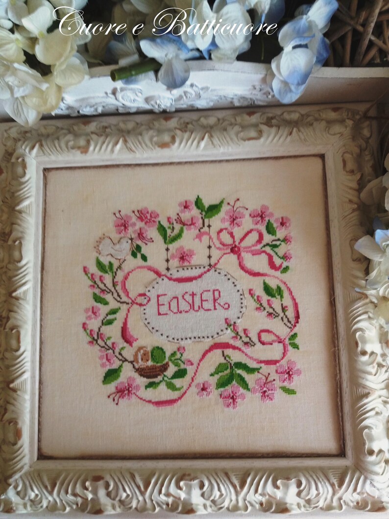 K&ouml;nnte beinhalten: Eine wei&szlig;e gerahmte Kreuzstich-Stickerei mit rosa Bl&uuml;ten, gr&uuml;nen Bl&auml;ttern und einem Band mit der Aufschrift "Easter".