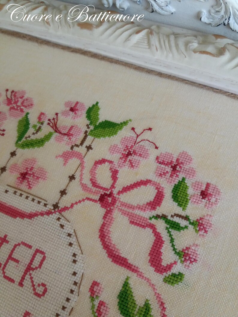 K&ouml;nnte beinhalten: Eine Kreuzstich-Stickerei mit einer rosa Schleife, rosa Blumen und gr&uuml;nen Bl&auml;ttern. Der Text "Sister" ist in einem wei&szlig;en ovalen Rahmen gestickt.