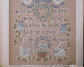 May include: A framed cross-stitch design featuring a blue and white alphabet with a floral wreath, two doves, a basket of Easter eggs, and a bunny. The design is on a light beige fabric and is framed in white.