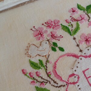 K&ouml;nnte beinhalten: Kreuzstich-Stickerei mit einem wei&szlig;en Vogel, rosa Blumen und gr&uuml;nen Bl&auml;ttern. Der Text "Cuore e Batticuore" ist oben im Design in braunem Garn gestickt.