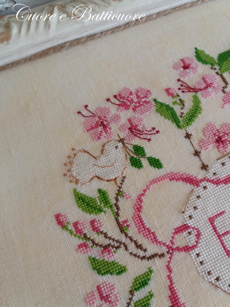K&ouml;nnte beinhalten: Kreuzstich-Stickerei mit einem wei&szlig;en Vogel, rosa Blumen und gr&uuml;nen Bl&auml;ttern. Der Text "Cuore e Batticuore" ist oben im Design in braunem Garn gestickt.