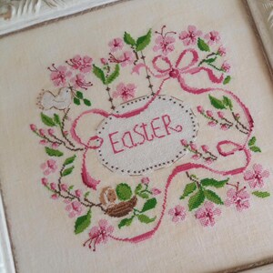 K&ouml;nnte beinhalten: Ein wei&szlig;er Stoff mit rosa und gr&uuml;ner Stickerei, die einen Vogel, ein Nest und ein Band mit dem Wort "Ostern" darauf darstellt.