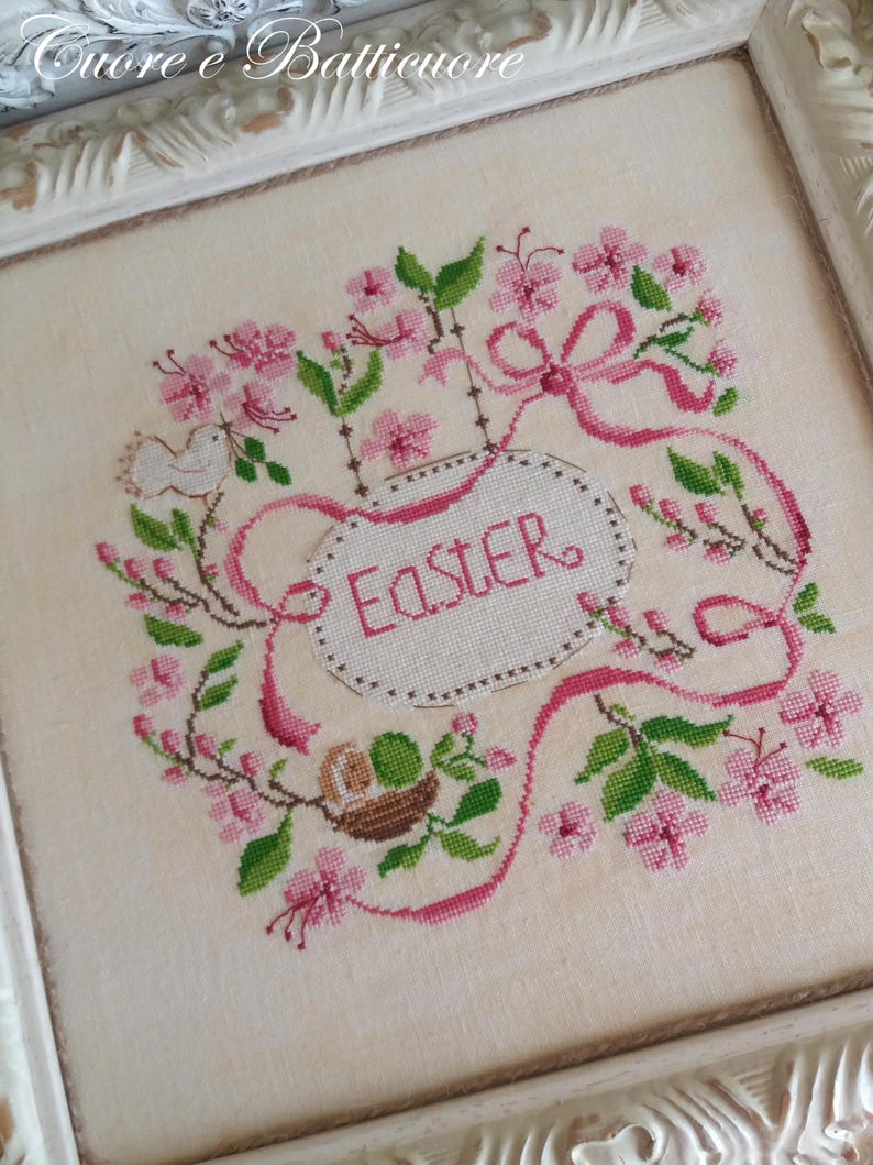 K&ouml;nnte beinhalten: Ein wei&szlig;er Stoff mit rosa und gr&uuml;ner Stickerei, die einen Vogel, ein Nest und ein Band mit dem Wort "Ostern" darauf darstellt.