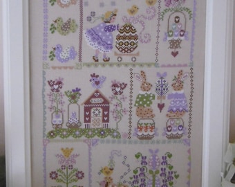 May include: Framed cross-stitch artwork featuring Easter-themed designs. The artwork includes a girl with an egg, bunnies, birds, and floral elements in pastel colors. The frame is white, and the overall style is quaint and decorative.