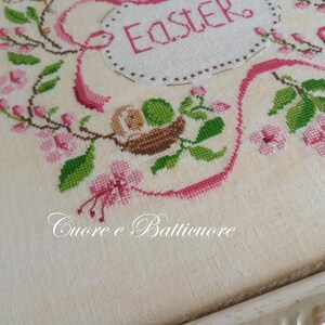 K&ouml;nnte beinhalten: Eine wei&szlig;e Kreuzstich-Stickerei mit rosa Blumen und gr&uuml;nen Bl&auml;ttern. Die Stickerei ist in einem wei&szlig;en Rahmen gerahmt. Der Text "Easter" ist in rosa auf der Stickerei gestickt.