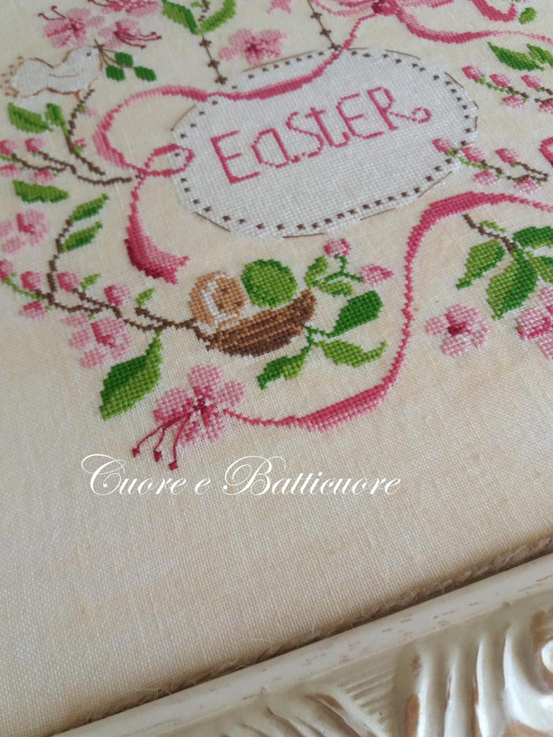 K&ouml;nnte beinhalten: Eine wei&szlig;e Kreuzstich-Stickerei mit rosa Blumen und gr&uuml;nen Bl&auml;ttern. Die Stickerei ist in einem wei&szlig;en Rahmen gerahmt. Der Text "Easter" ist in rosa auf der Stickerei gestickt.