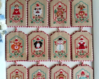 May include: Twelve cross-stitch ornaments with a Christmas theme. Each ornament features a different design, including a snowman, a penguin, a reindeer, a gingerbread man, and a Santa Claus. The ornaments are all beige with red and green accents. The text on the ornaments reads "Per Te", "For You", "Four To", and "Für Dich".