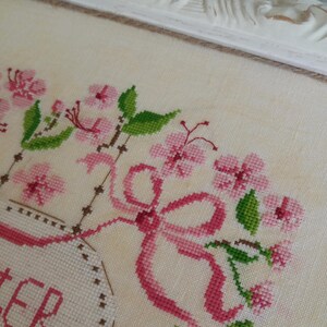 K&ouml;nnte beinhalten: Eine Kreuzstich-Stickerei mit einer rosa Schleife, rosa Blumen und gr&uuml;nen Bl&auml;ttern. Der Text "Sister" ist in einem wei&szlig;en ovalen Rahmen gestickt.