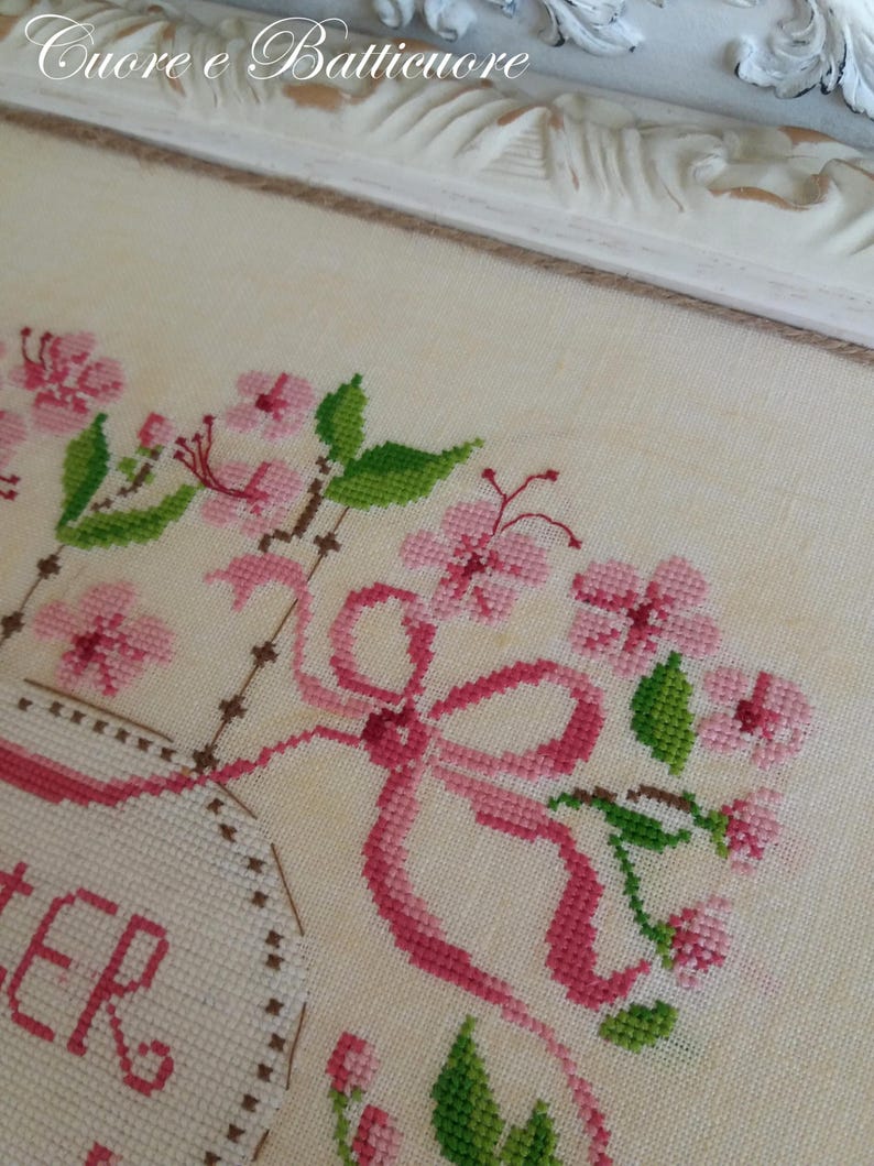 K&ouml;nnte beinhalten: Eine Kreuzstich-Stickerei mit einer rosa Schleife, rosa Blumen und gr&uuml;nen Bl&auml;ttern. Der Text "Sister" ist in einem wei&szlig;en ovalen Rahmen gestickt.