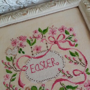 K&ouml;nnte beinhalten: Eine Kreuzstich-Stickerei mit einem rosa und gr&uuml;nen Blumendesign und einem wei&szlig;en Oval in der Mitte, auf dem "Easter" steht.