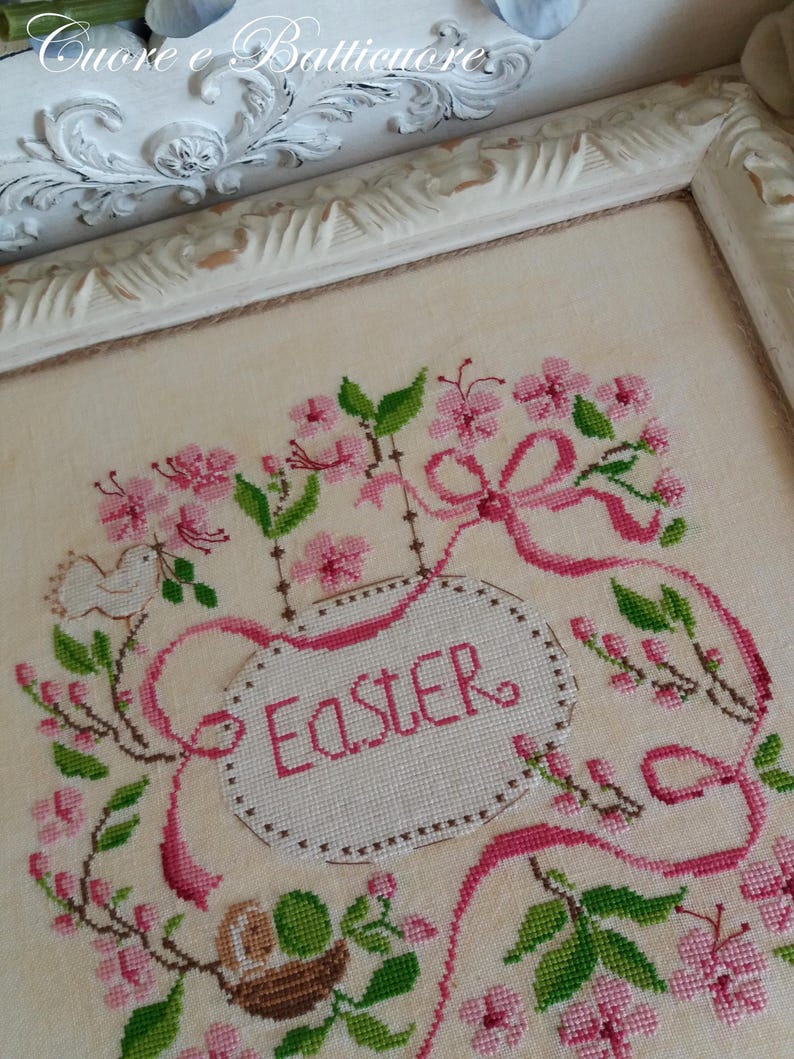 K&ouml;nnte beinhalten: Eine Kreuzstich-Stickerei mit einem rosa und gr&uuml;nen Blumendesign und einem wei&szlig;en Oval in der Mitte, auf dem "Easter" steht.