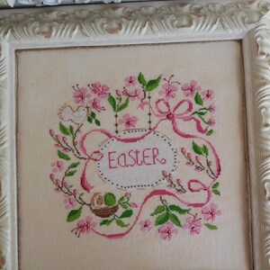 K&ouml;nnte beinhalten: Eine gerahmte Kreuzstich-Stickerei mit einem rosa und gr&uuml;nen Blumendesign, einem wei&szlig;en Vogel und dem Wort "Easter" in einer rosa Ellipse.