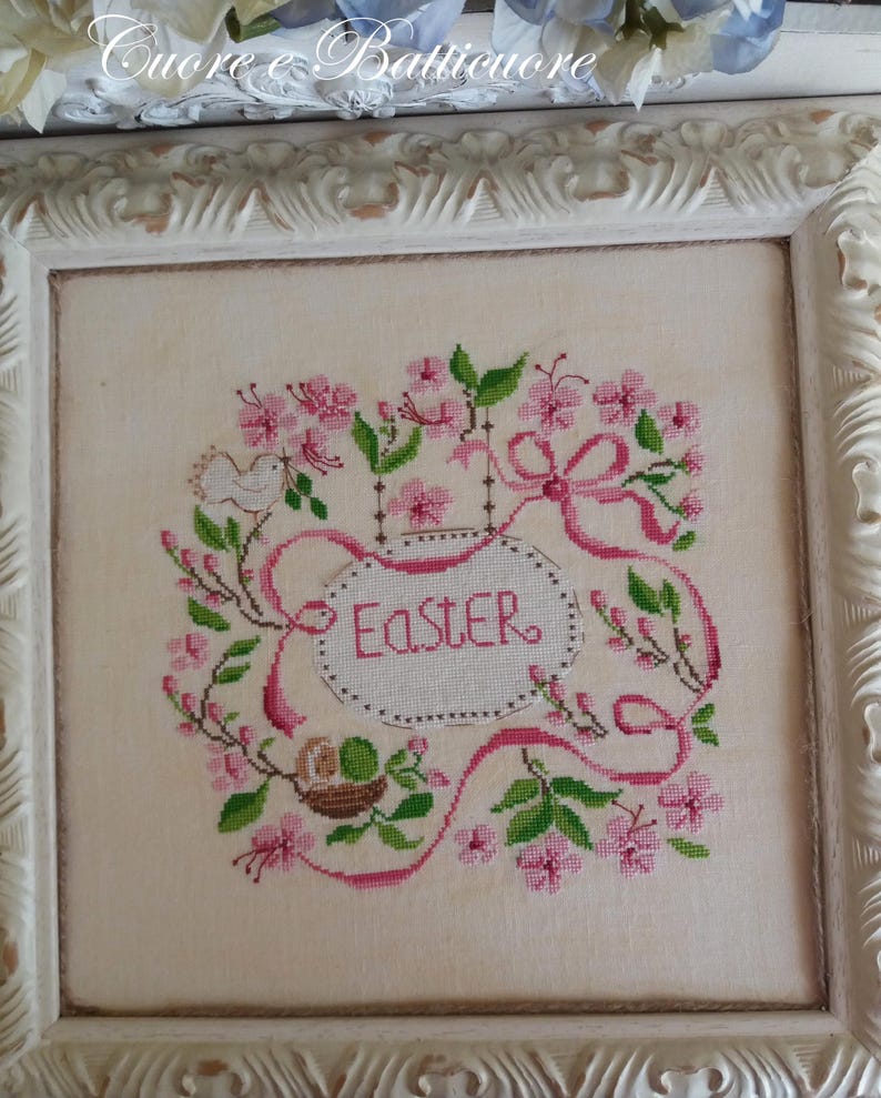 K&ouml;nnte beinhalten: Eine gerahmte Kreuzstich-Stickerei mit einem rosa und gr&uuml;nen Blumendesign, einem wei&szlig;en Vogel und dem Wort "Easter" in einer rosa Ellipse.