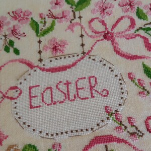 K&ouml;nnte beinhalten: Eine wei&szlig;e Kreuzstich-Stickerei mit rosa und gr&uuml;nen Blumen und dem Wort "Easter" in einem rosa Oval.