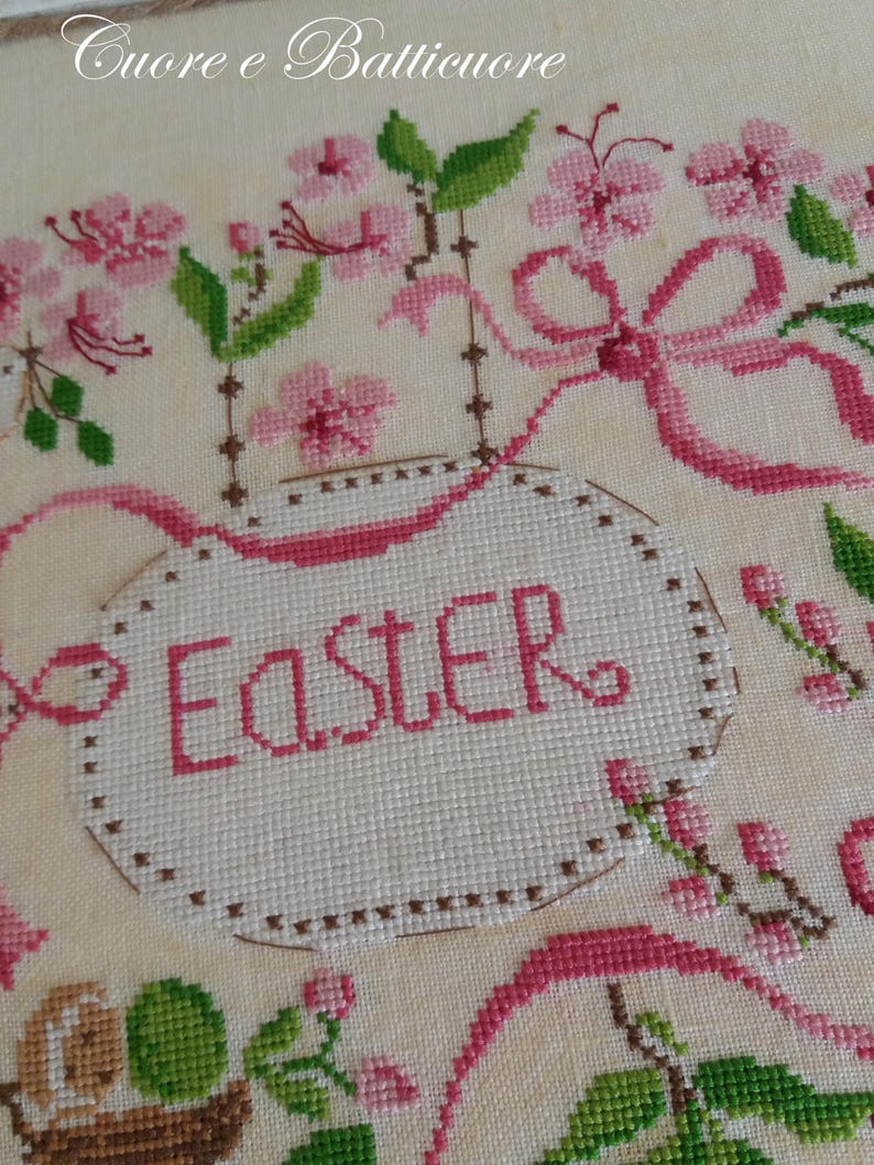 K&ouml;nnte beinhalten: Eine wei&szlig;e Kreuzstich-Stickerei mit rosa und gr&uuml;nen Blumen und dem Wort "Easter" in einem rosa Oval.