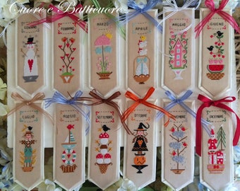 May include: Twelve wooden bookmarks with cross-stitch designs representing each month of the year. The bookmarks are decorated with ribbons in various colors and feature Italian month names.