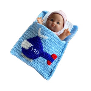 Puede incluir: Un saco de dormir para bebé de crochet azul con un diseño de helicóptero y el número 110 bordado en él. Una muñeca está acostada dentro del saco de dormir.