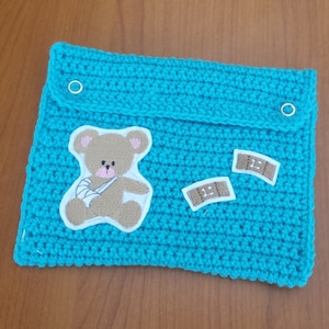 Pode incluir: Uma bolsa de crochê turquesa com uma aba presa por dois ilhós prateados. A bolsa apresenta um ursinho de pelúcia aplicado com um braço enfaixado e duas pequenas aplicações de curativos.