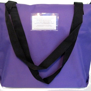 May include: A purple tote bag with black straps. The bag has a rectangular clear pocket with white text. The bag has a zipper closure at the top. The straps are sewn to the bag and extend down the sides.