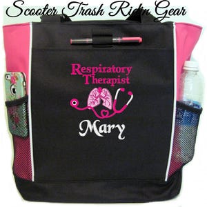 May include: Black tote bag with pink accents and a white zipper. The bag has a mesh pocket on the side and a zippered pocket on the front. The bag is embroidered with the text "Respiratory Therapist" and a stethoscope with lungs. The name "Mary" is embroidered below the stethoscope.