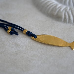 May include: A necklace featuring a gold-toned, hammered metal fish pendant attached to a dark blue cord. The cord includes small, gold-toned metal beads and is knotted near the pendant, which has a smooth, reflective surface and a simple tail fin.