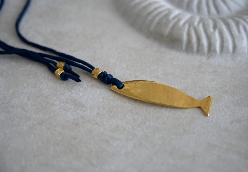 May include: A necklace featuring a gold-toned, hammered metal fish pendant attached to a dark blue cord. The cord includes small, gold-toned metal beads and is knotted near the pendant, which has a smooth, reflective surface and a simple tail fin.