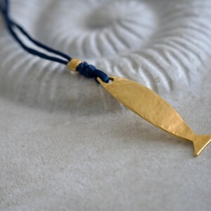 May include: A necklace featuring a gold-toned, hammered metal fish pendant suspended from a dark blue cord. The pendant is attached to the cord with a small gold-toned bead and a knot, creating a simple, clean design for this piece of jewelry.