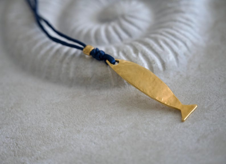 May include: A necklace featuring a gold-toned, hammered metal fish pendant suspended from a dark blue cord. The pendant is attached to the cord with a small gold-toned bead and a knot, creating a simple, clean design for this piece of jewelry.