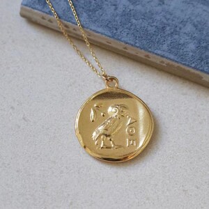 Op de afbeelding: Goudkleurige hangerketting met een ronde hanger met een reli&euml;f van een uil en olijftak. De ketting heeft een fijne gouden ketting. De hanger ligt op een lichte ondergrond met een blauwe achtergrond.