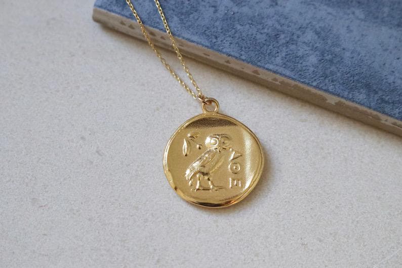 Op de afbeelding: Goudkleurige hangerketting met een ronde hanger met een reli&euml;f van een uil en olijftak. De ketting heeft een fijne gouden ketting. De hanger ligt op een lichte ondergrond met een blauwe achtergrond.