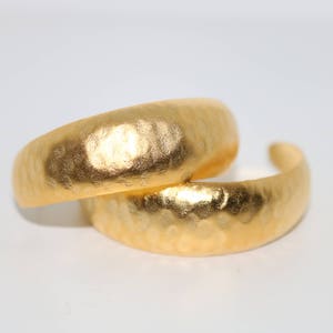 May include: Two gold-toned hammered metal hoop earrings. The earrings are thick and have a rounded shape.