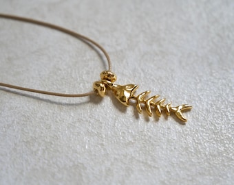May include: A gold-toned fish skeleton pendant suspended from a thin, light beige cord. The pendant features a textured, metallic finish with a detailed spinal structure and a small, rounded bead connector at the top of the fish head.