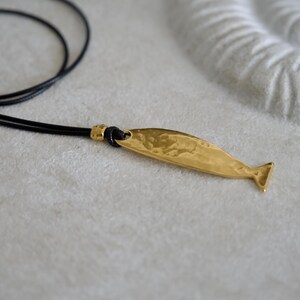 May include: A gold-toned metal pendant shaped like a whale, featuring a textured, hammered surface. The pendant is attached to a thin, black cord necklace by a small gold-toned metal bead, creating a simple and distinct piece of jewelry.