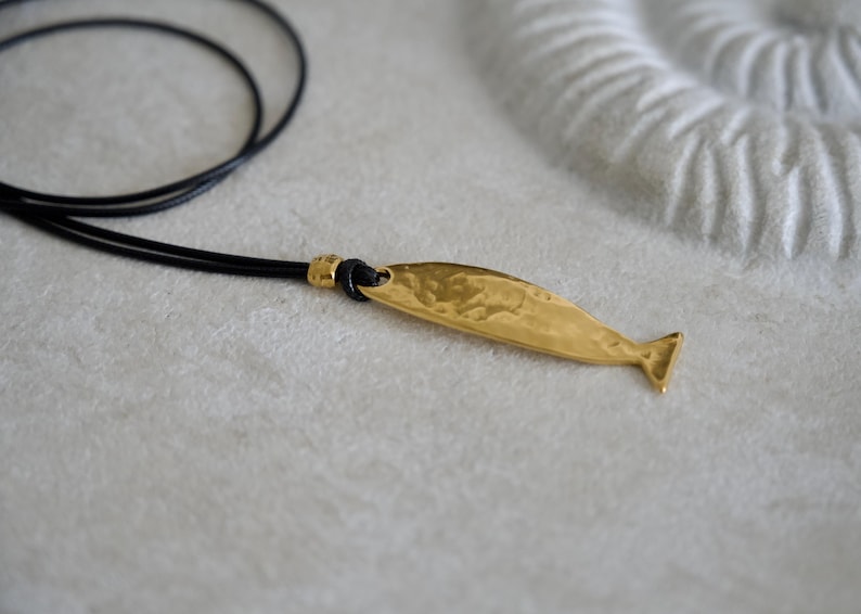 May include: A gold-toned metal pendant shaped like a whale, featuring a textured, hammered surface. The pendant is attached to a thin, black cord necklace by a small gold-toned metal bead, creating a simple and distinct piece of jewelry.