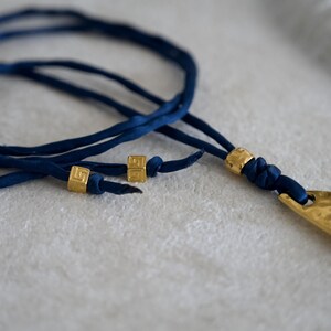 May include: A necklace featuring a long, hammered gold-tone metal pendant attached to a dark blue fabric cord. The cord is accented with three small, square gold-tone beads, two of which are positioned on the loose ends of the cord.
