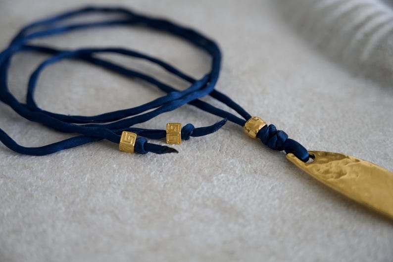 May include: A necklace featuring a long, hammered gold-tone metal pendant attached to a dark blue fabric cord. The cord is accented with three small, square gold-tone beads, two of which are positioned on the loose ends of the cord.