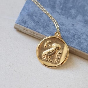 Op de afbeelding: Goudkleurige hangerketting met een cirkelvormig muntontwerp met een uil en een olijftak. De hanger hangt aan een delicate gouden ketting. De ketting ligt op een blauw oppervlak.
