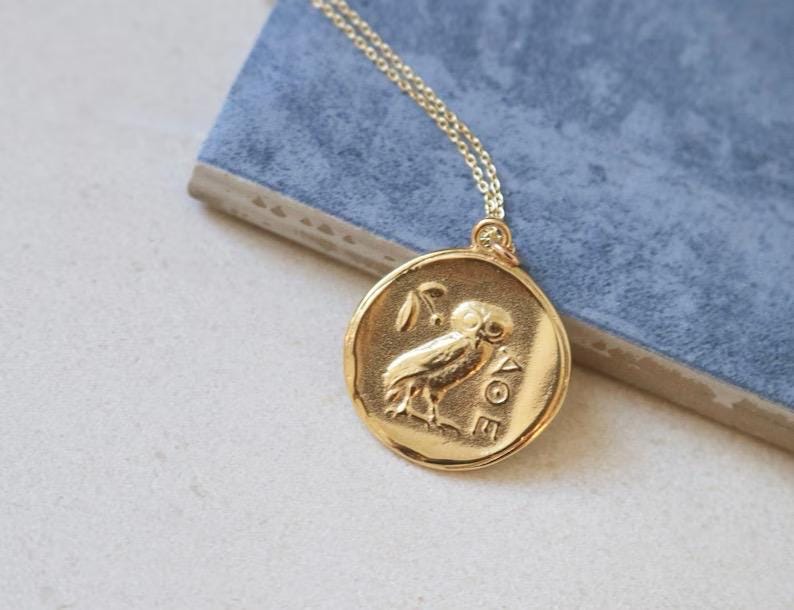 Op de afbeelding: Goudkleurige hangerketting met een cirkelvormig muntontwerp met een uil en een olijftak. De hanger hangt aan een delicate gouden ketting. De ketting ligt op een blauw oppervlak.