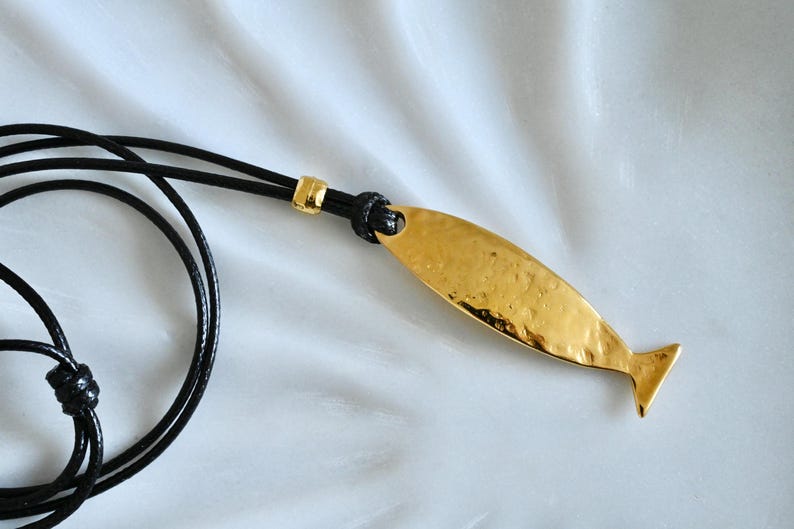 May include: A gold-toned, hammered-finish fish pendant suspended from a black cord necklace. The pendant features an elongated, streamlined shape with a distinct tail fin, attached to the cord by a small gold-toned metal bead and a knot.