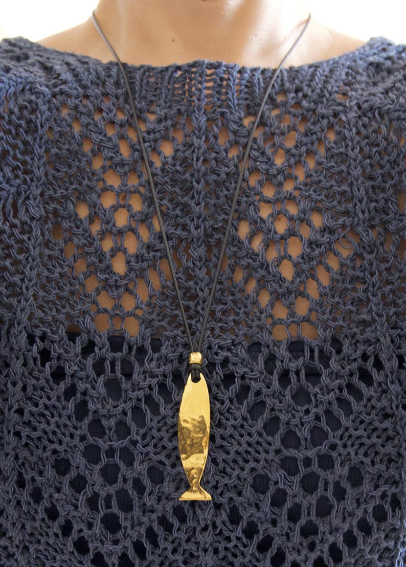 May include: A gold-toned, hammered metal fish pendant suspended from a thin black cord. The pendant features a smooth, reflective surface with a textured, dimpled finish, hanging vertically from a small gold bead at the top of the cord.