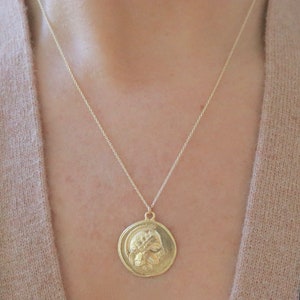 Op de afbeelding: Een goudkleurige hangerketting met een cirkelvormige, muntachtige bedel met een gedetailleerd reli&euml;f van een klassieke figuur. De hanger hangt aan een fijne ketting. De ketting wordt gedragen over een trui in neutrale tinten.