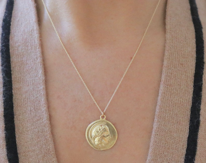 Op de afbeelding: Een goudkleurige hangerketting met een cirkelvormige, muntachtige bedel met een gedetailleerd reli&euml;f van een klassieke figuur. De hanger hangt aan een fijne ketting. De ketting wordt gedragen over een trui in neutrale tinten.