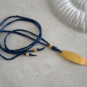 May include: A necklace featuring a gold-toned, hammered metal fish pendant suspended from a dark blue cord. The cord is accented with three small, square gold-toned beads, two of which are positioned near the ends and one near the pendant.