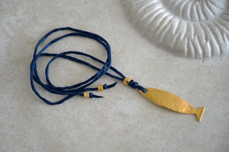 May include: A necklace featuring a gold-toned, hammered metal fish pendant suspended from a dark blue cord. The cord is accented with three small, square gold-toned beads, two of which are positioned near the ends and one near the pendant.