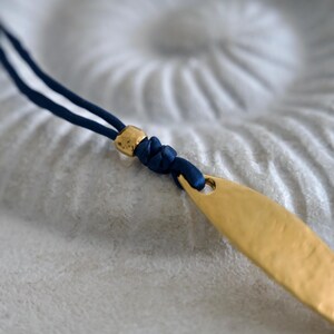 May include: A necklace featuring a dark blue cord, a small metallic gold bead, and a textured, elongated gold-toned pendant. The cord is knotted securely through the top of the pendant, creating a simple design with contrasting blue and gold colors.